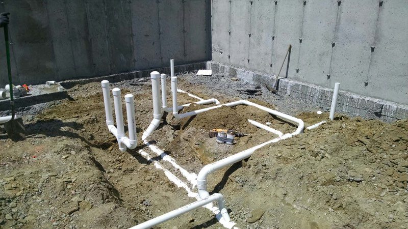 PVC plumbing rough-in in a new residential foundation: white drain and vent pipes laid in trenches on compacted soil against concrete foundation walls, showing bathroom/kitchen branch stacks and waste lines ready for backfill.