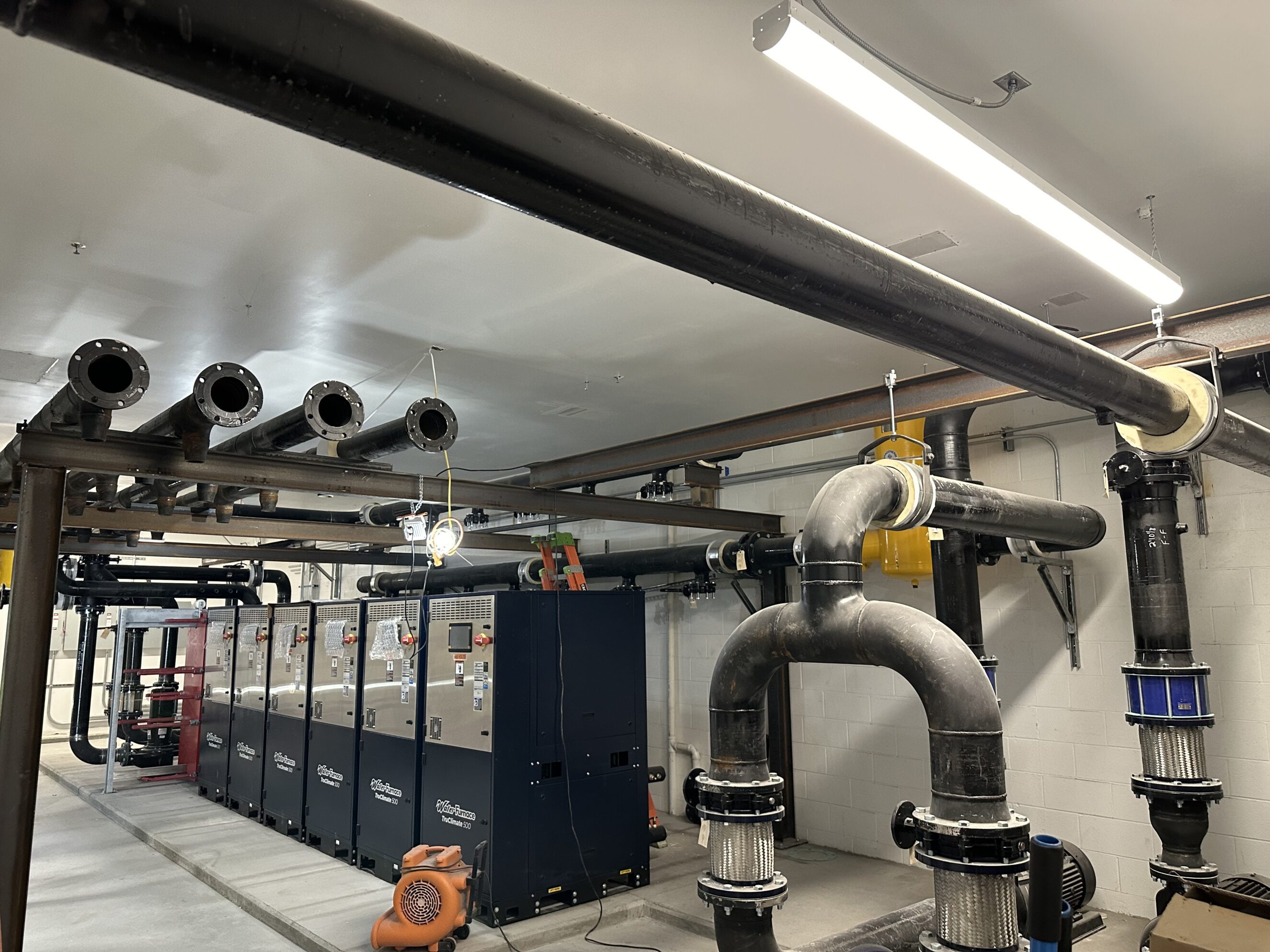 Large commercial mechanical room with black overhead and floor-mounted industrial piping, large U‑shaped drainage and insulated joints, a row of blue commercial boilers/chillers, conduit and hanging supports, and maintenance ladder and blower present—commercial plumbing installation in a utility room.