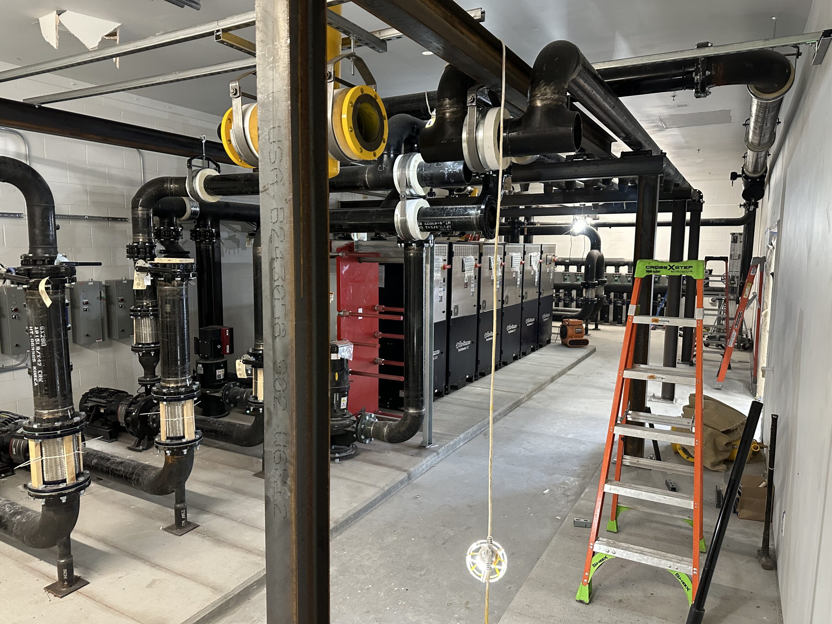 Commercial mechanical room under construction with black industrial plumbing and large looped pipes, pumps and control panels along the left, red boiler/heat exchanger units center-left, orange step ladder and temporary work light hanging from ceiling on the right, concrete floor and white block walls